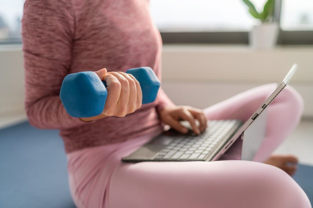 Fitness workout and working on laptop computer - woman multitasking remote work at home indoors lifting dumbbell weights for strength training while inside | ThirstyAffiliates Best Affiliate Programs For Health and Wellness